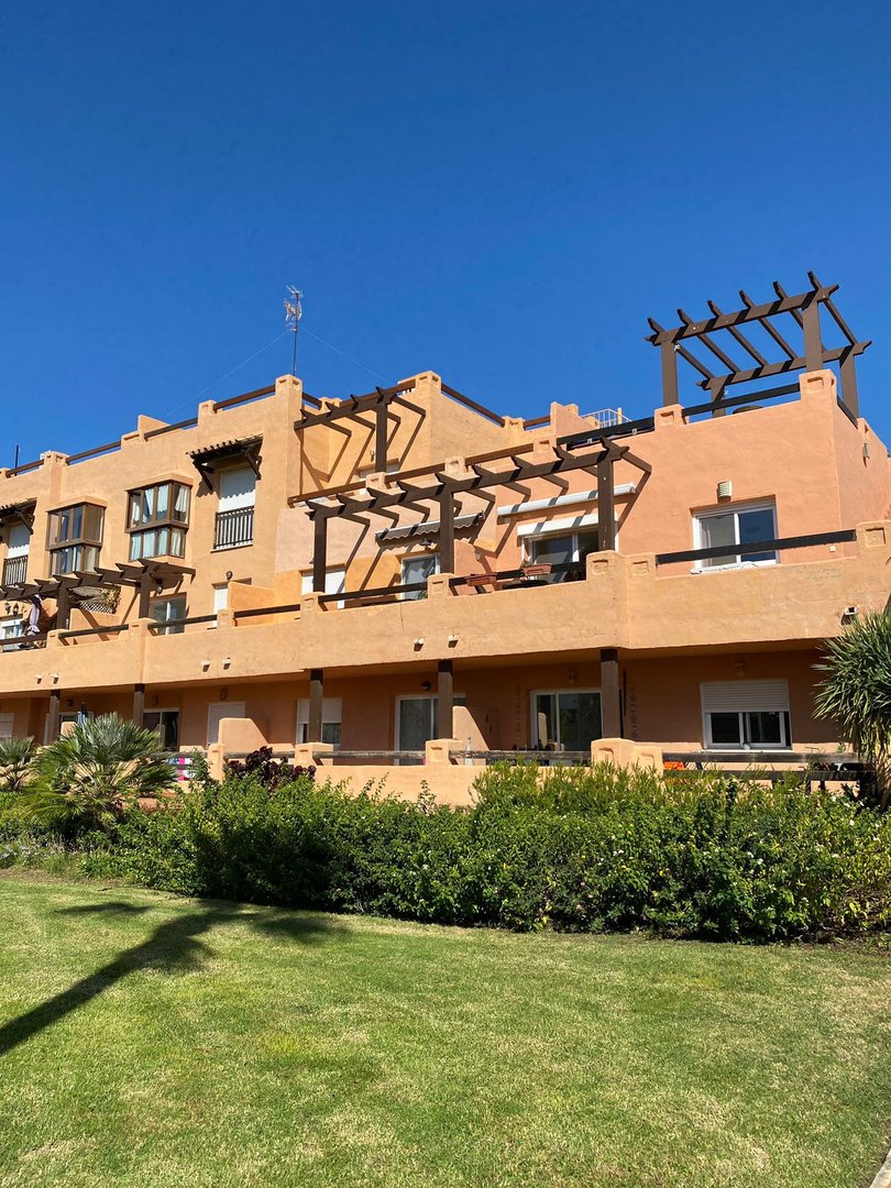 Apartment building facade with terraces and pergolas, overlooking a green lawn and bushes under a clear blue sky.