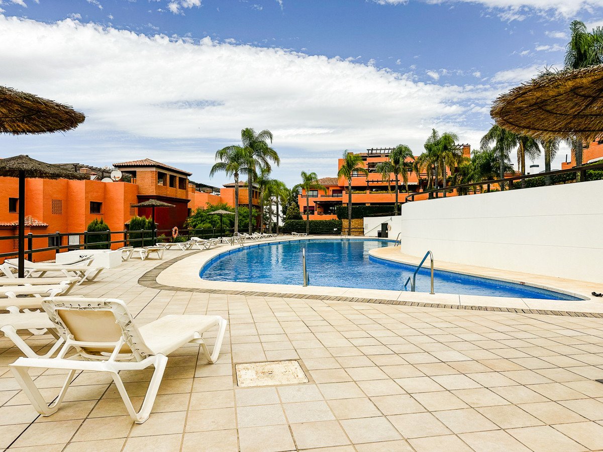 Resort apartments with a large, curved swimming pool surrounded by loungers and palm trees under a blue sky with clouds.