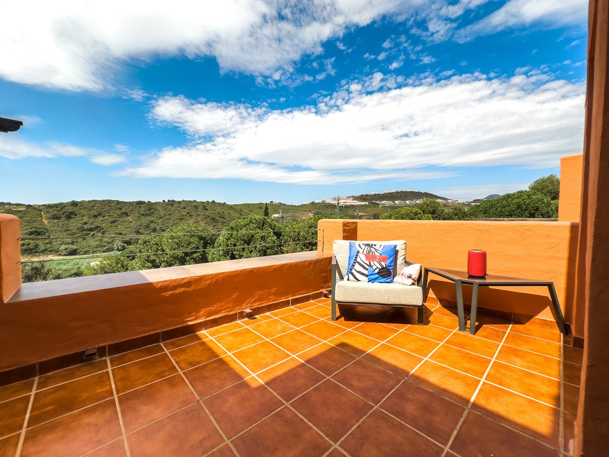 Terrace with seating and a side table overlooking a lush green mountain landscape under a bright blue sky with clouds.