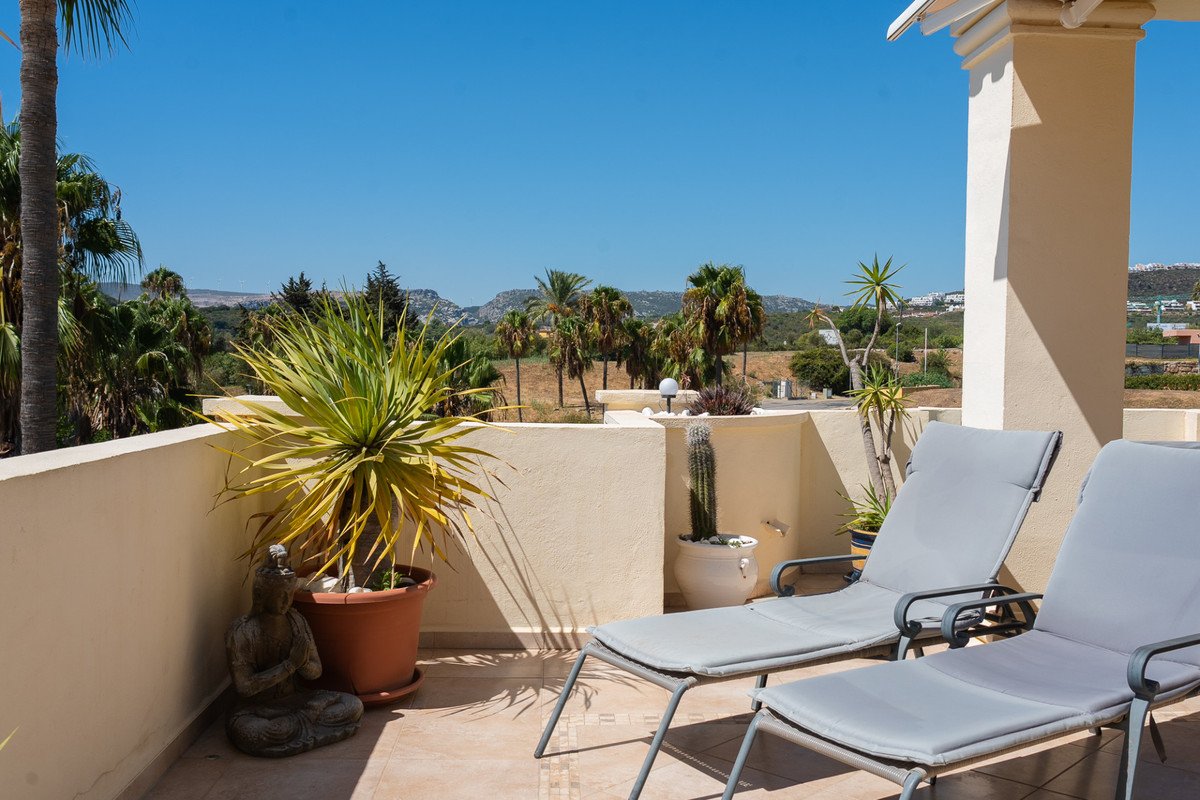 A sunny terrace with two lounge chairs, potted plants, and a view of distant mountains and palm trees.