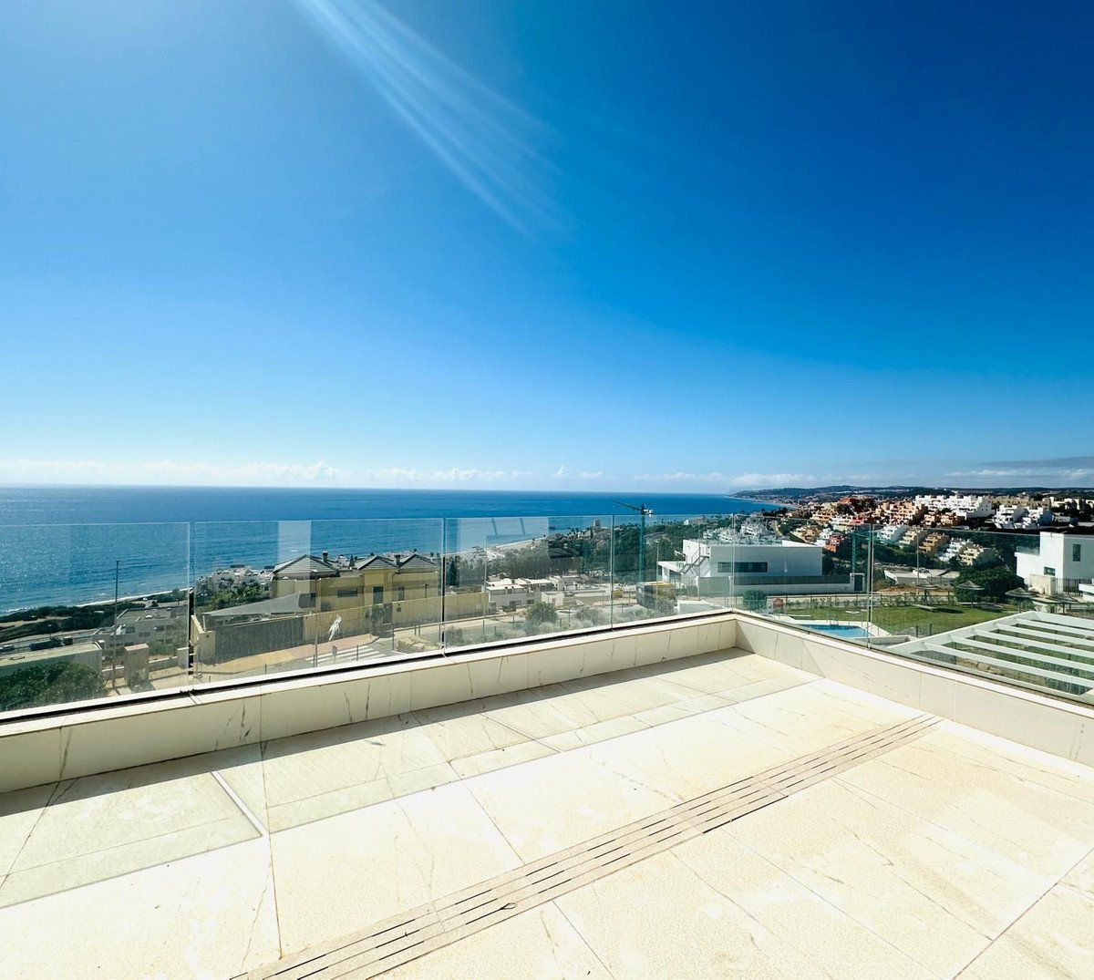 Spacious terrace with glass railings offers a panoramic view of the sea and a modern residential complex under a clear blue sky.