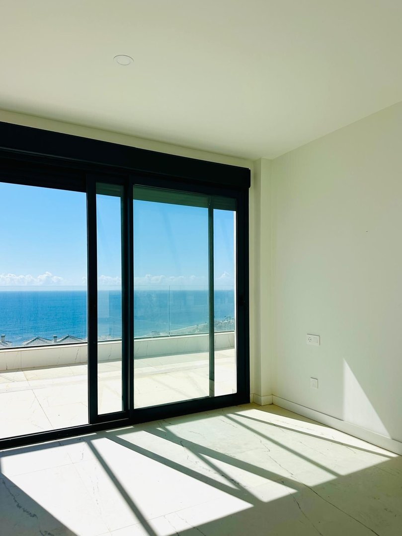 Empty room with large sliding glass doors opening to a balcony with a stunning sea view under a clear blue sky.