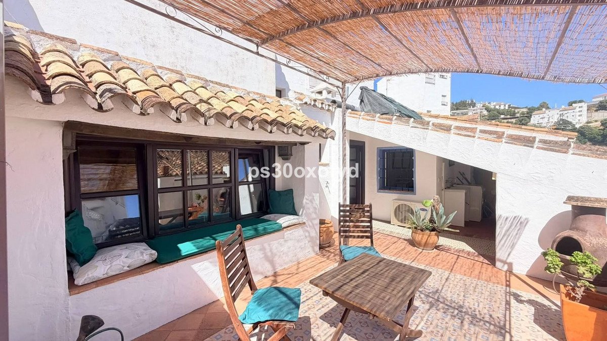 Terrace with wooden chairs, table, and cushioned bench seating. Overlooks a charming Mediterranean town with hills in the background.