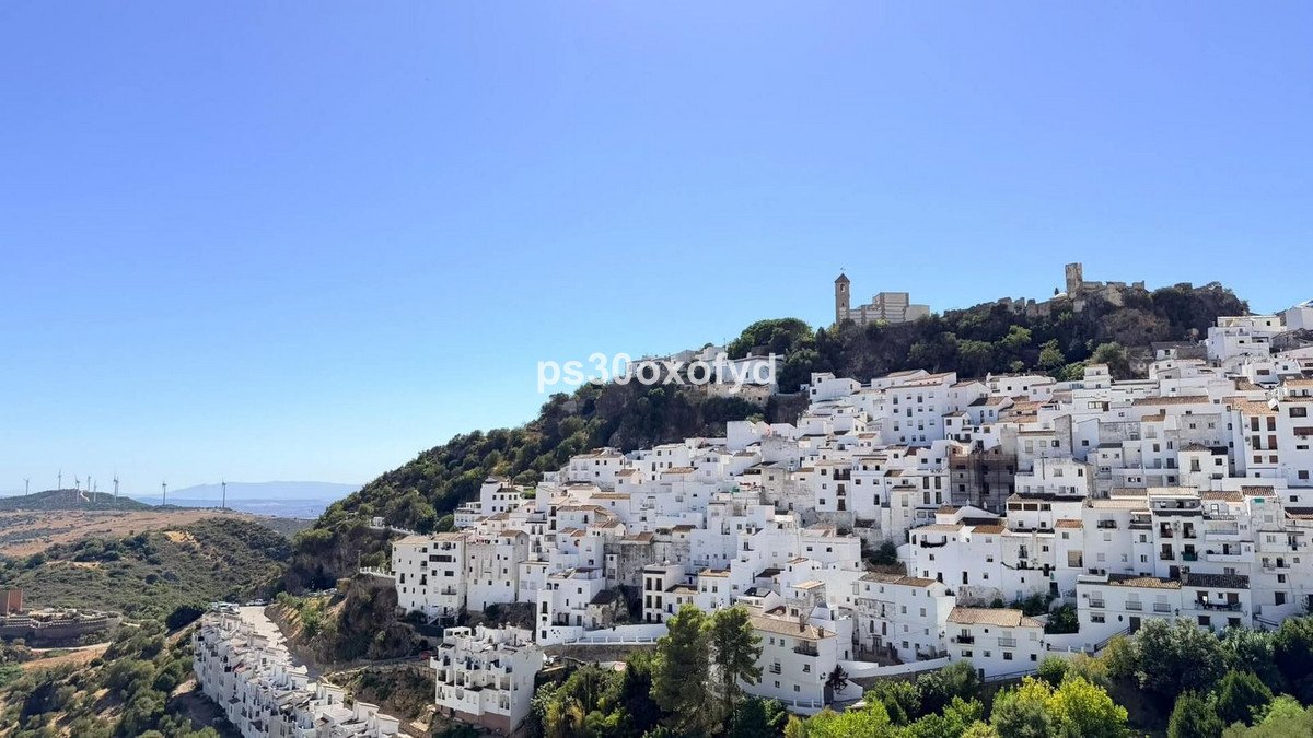 A picturesque hillside village with white buildings, featuring a castle ruin on the hill and distant windmills under a clear blue sky.