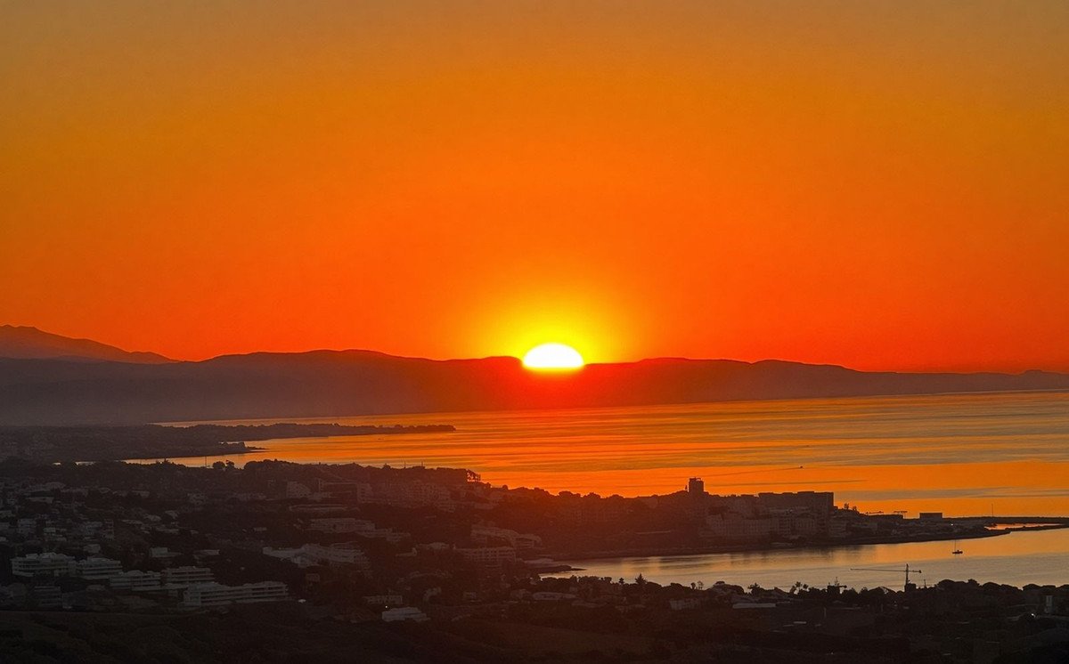 A vibrant sunset over a coastal town and the calm sea, with mountains in the distance.