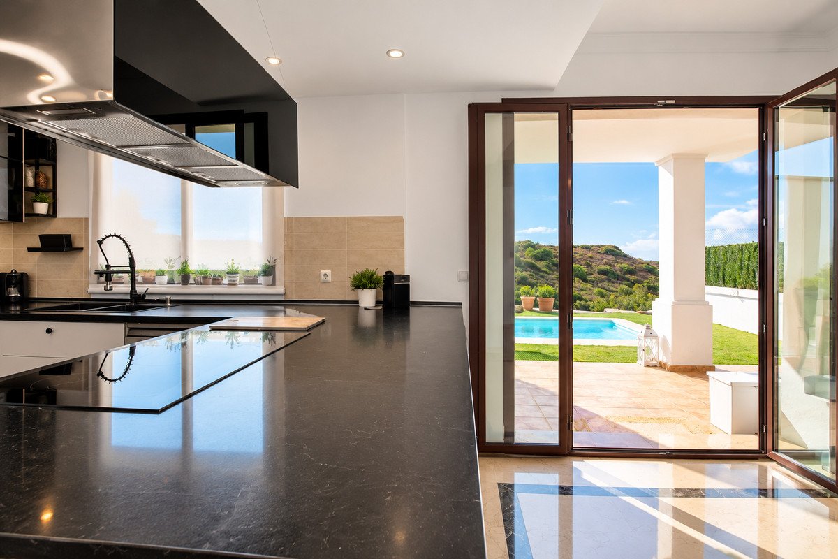 Modern kitchen with dark countertops opens to a backyard with a swimming pool and lush green hills under a blue sky.