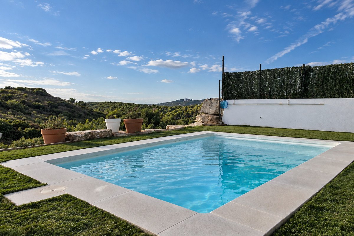 A rectangular swimming pool with clear blue water, surrounded by green grass and potted plants, with a view of rolling hills under a blue sky.