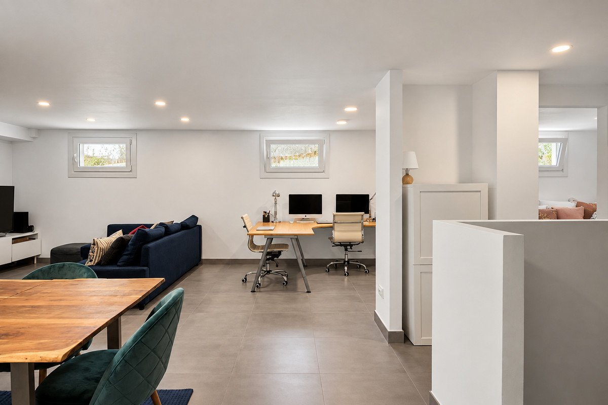Modern living space with a home office setup featuring dual monitors and ergonomic chairs, next to a blue sofa and wooden dining table.