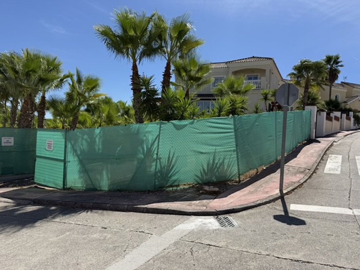 Construction site with green mesh fencing in front of a villa with palm trees, on a sunny day.