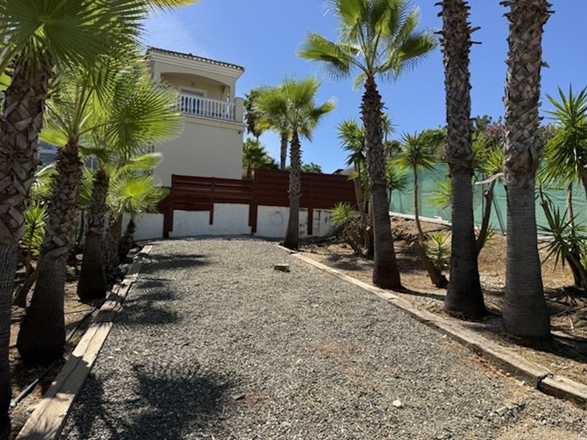 Gravel driveway lined with palm trees leading to a villa with a balcony under a clear blue sky.