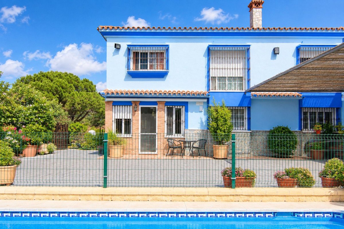 A light blue two-story house with a tiled roof, a swimming pool, and a lush garden with trees and flowers.