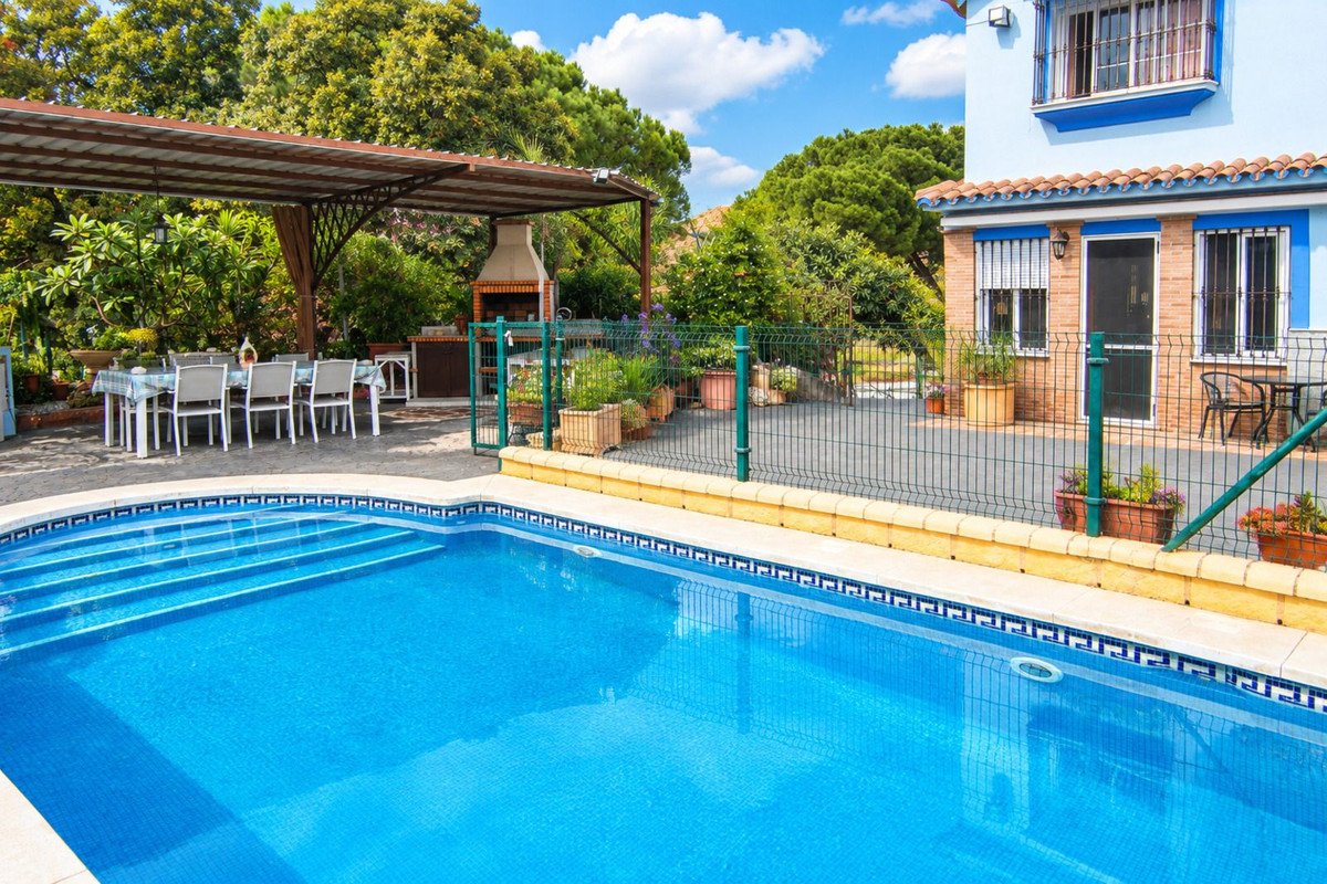 Outdoor swimming pool with steps, covered dining area with table and chairs, and a brick barbecue grill on a sunny day.