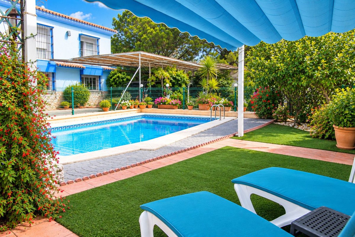 A bright blue swimming pool and lush garden with sunbeds and a blue awning, surrounded by flowering plants and trees.