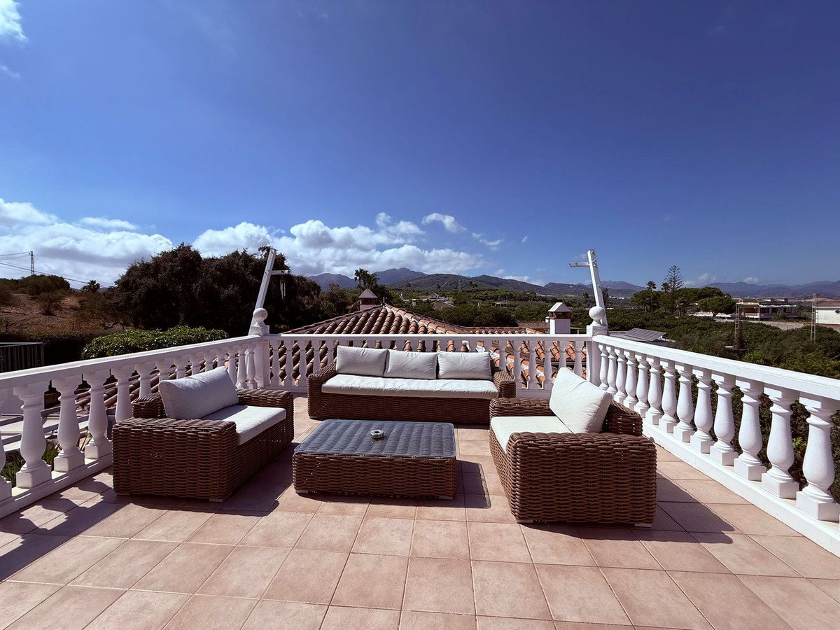 Rooftop terrace with wicker furniture, white balustrade, and mountain views under a clear blue sky.