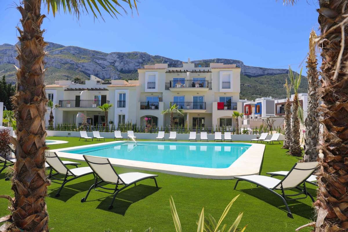 Luxury apartment building with a swimming pool and sun loungers, overlooking mountains on a sunny day.