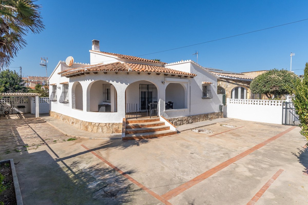 Exterior view of a white villa with arched covered terrace, stone base, tiled roof, and paved parking area.