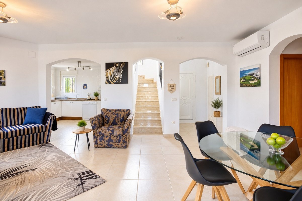 Open plan living, dining, and kitchen area with striped sofa, patterned armchair, glass table, and stairs in the background.