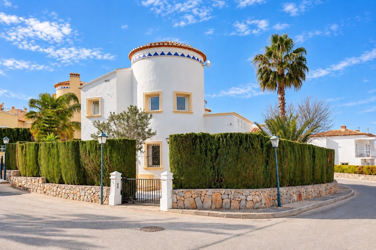 A white villa with a circular tower, surrounded by tall green hedges and a palm tree, under a blue sky.