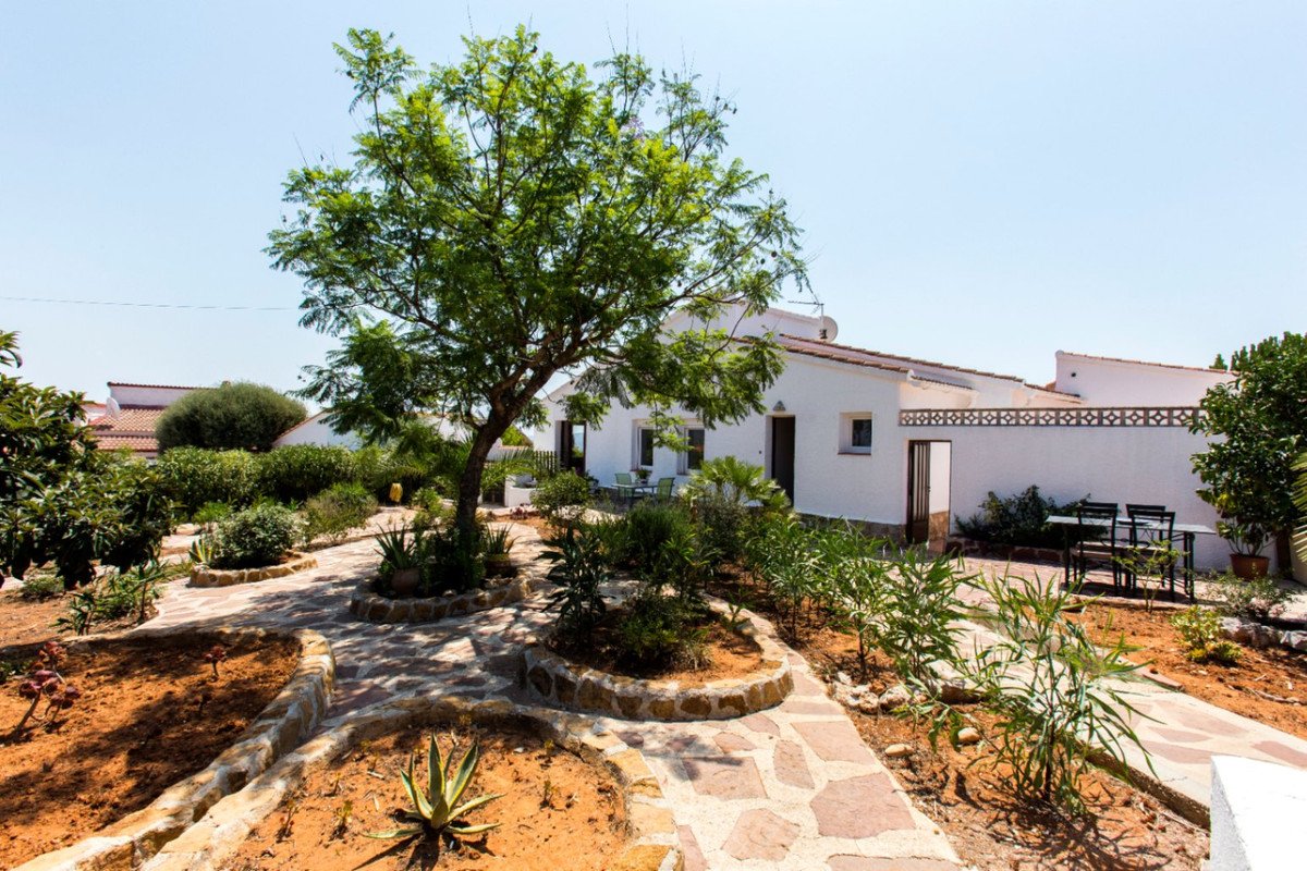 White villa with a lush garden, stone pathways, and a patio seating area under a large tree.