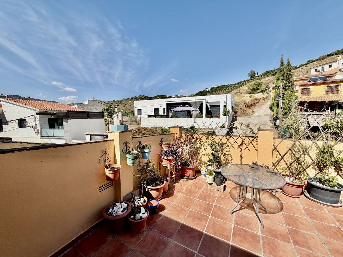 A sunny terrace with terracotta tiles, potted plants, a glass table, and views of houses and hills.