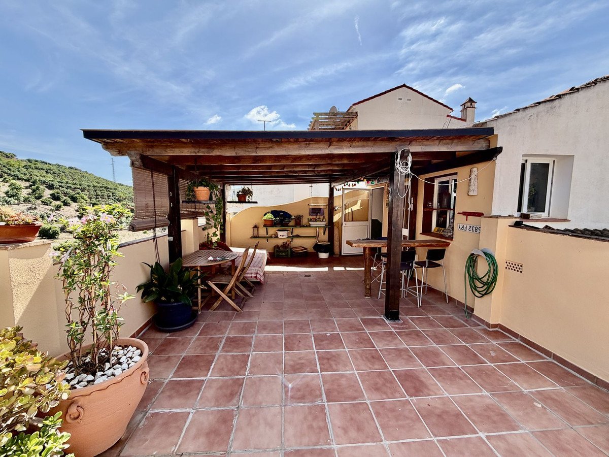 A covered terrace with terracotta flooring, featuring dining tables, bar seating, plants, and a view of a hillside under a blue sky.