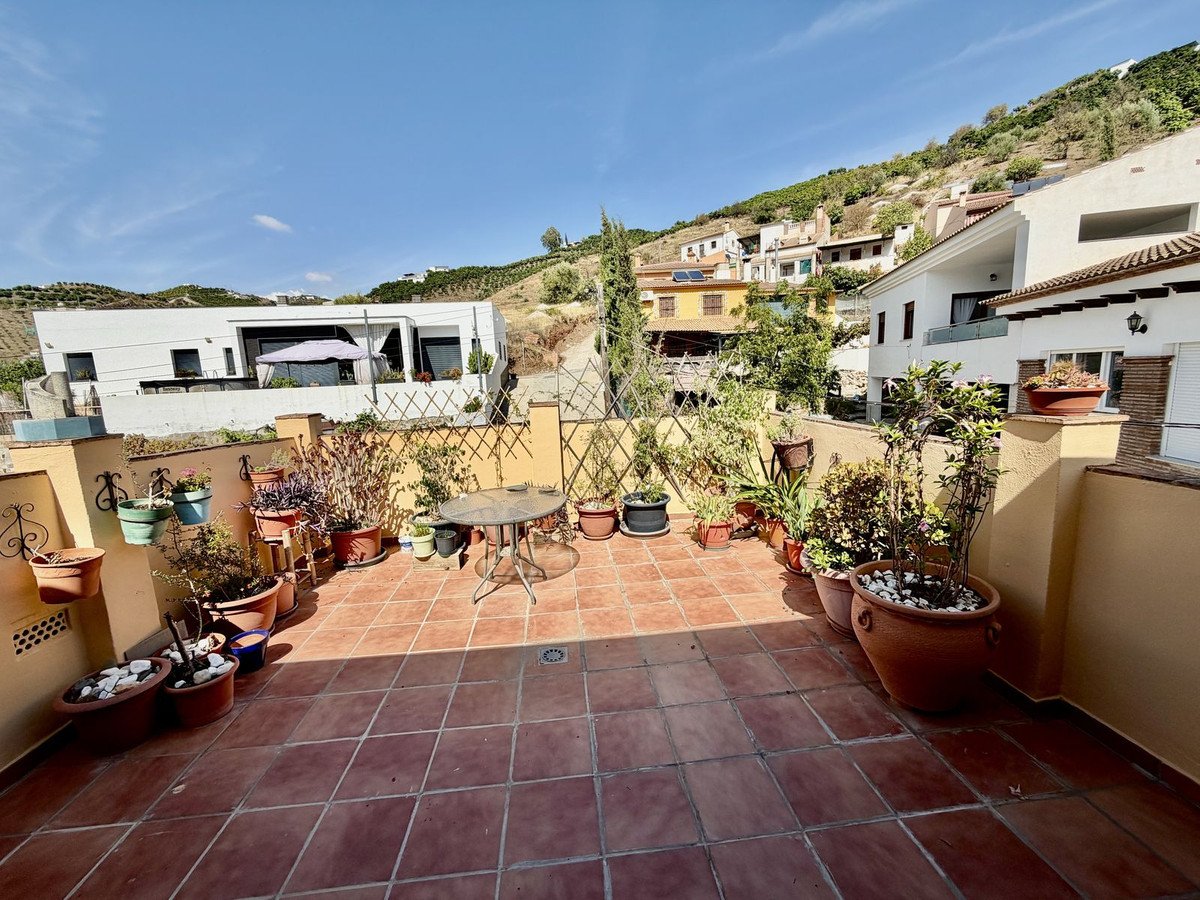 A sunny terrace with potted plants and a small table, overlooking houses and a hillside.