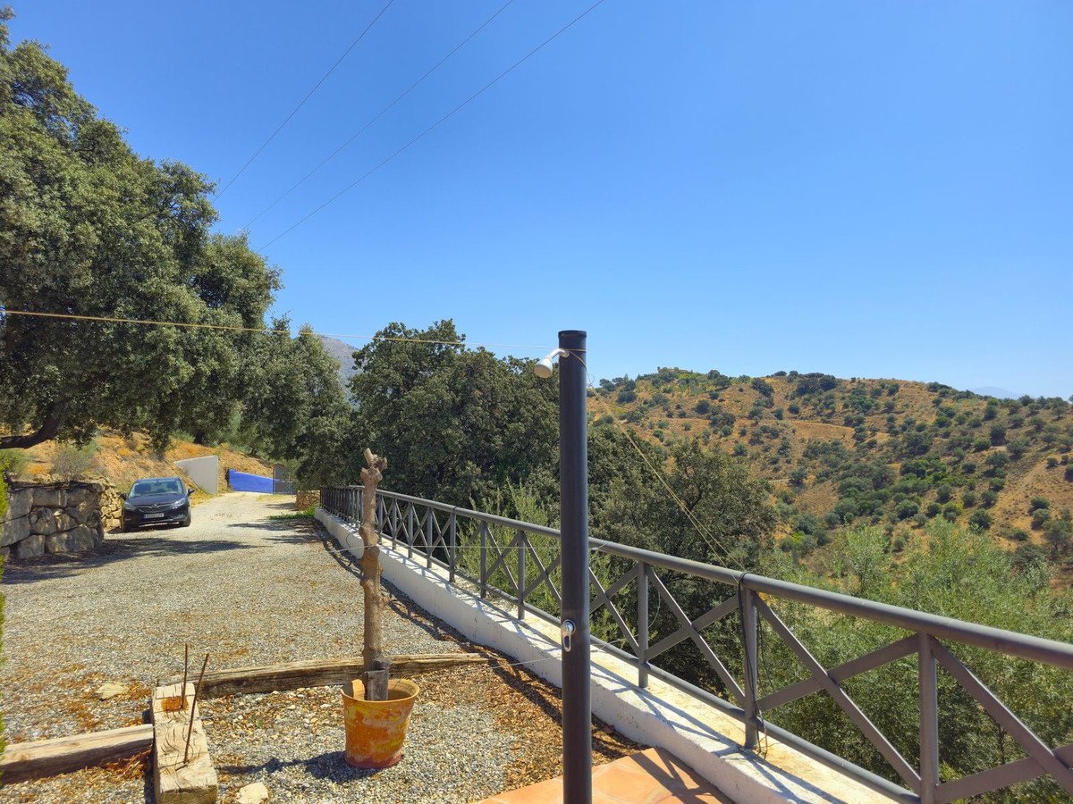 Outdoor terrace with a railing overlooking a sunny, hilly landscape with trees and a clear blue sky.