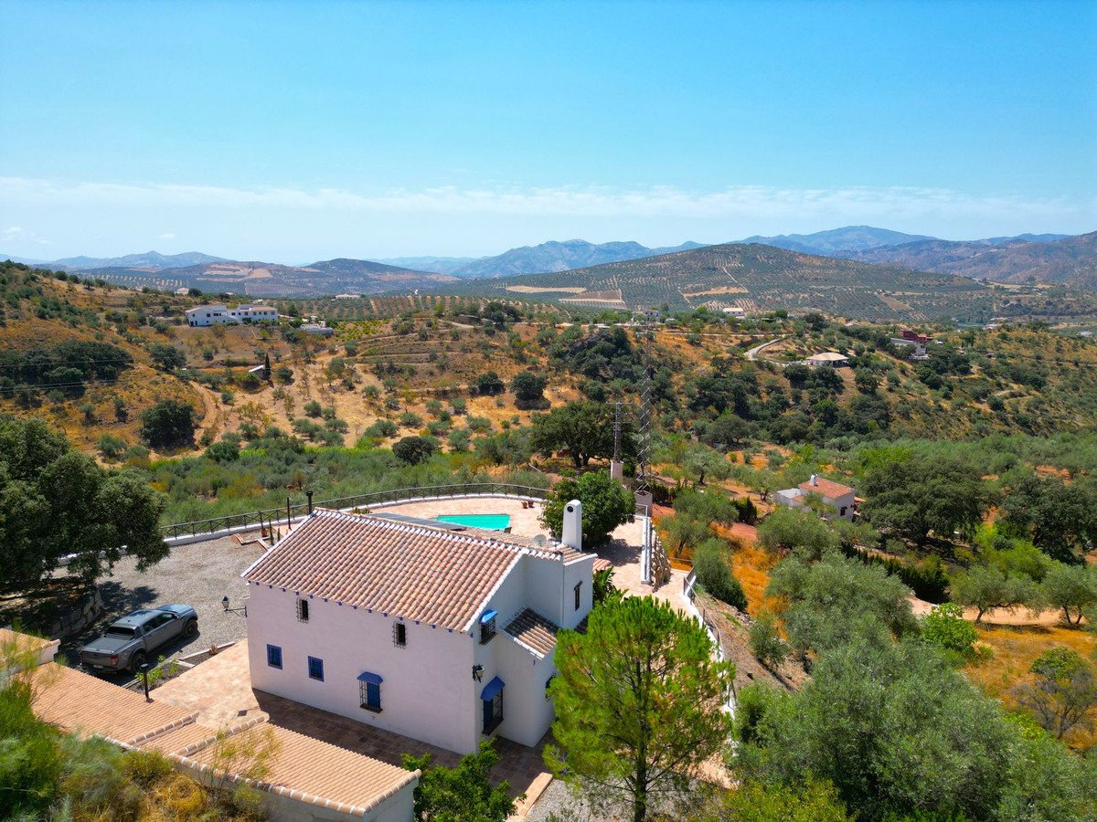 A white villa with blue shutters and a swimming pool, set in rolling hills with olive groves and distant mountains.