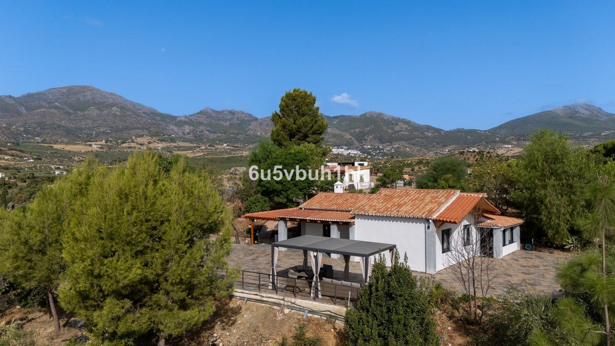A white villa with an orange tiled roof and a covered patio, set against a backdrop of lush green trees and rolling mountains under a clear blue sky.