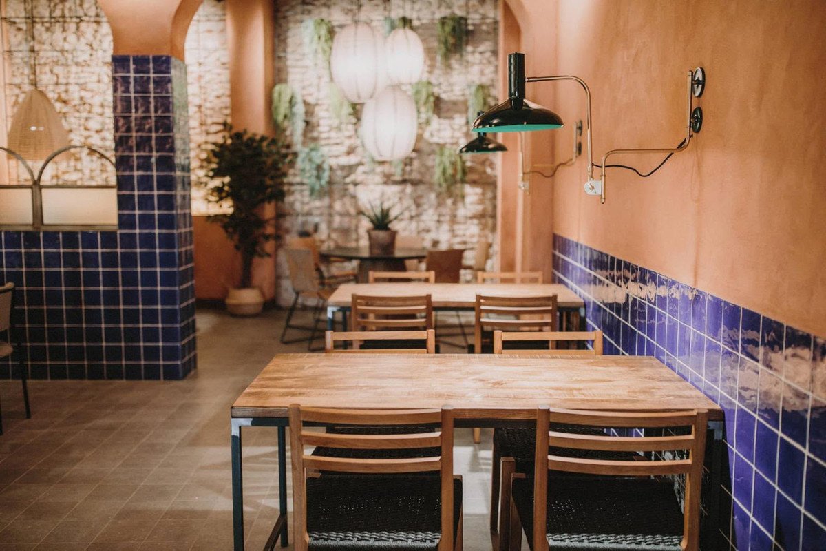 Restaurant interior with wooden tables, chairs, blue tiled walls, and hanging pendant lights.