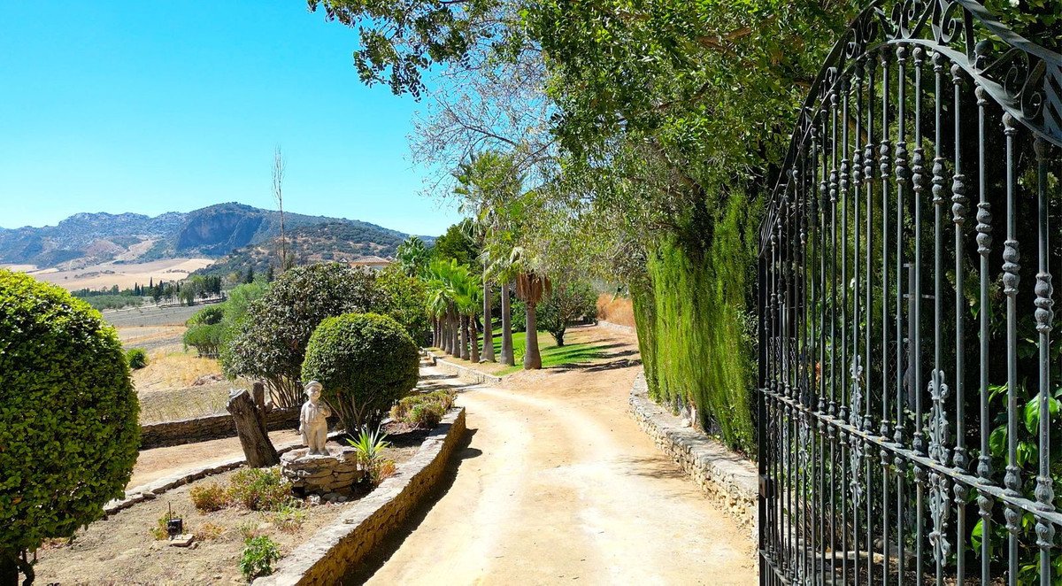 A gated driveway leads to an estate, with manicured gardens and distant mountains under a clear blue sky.