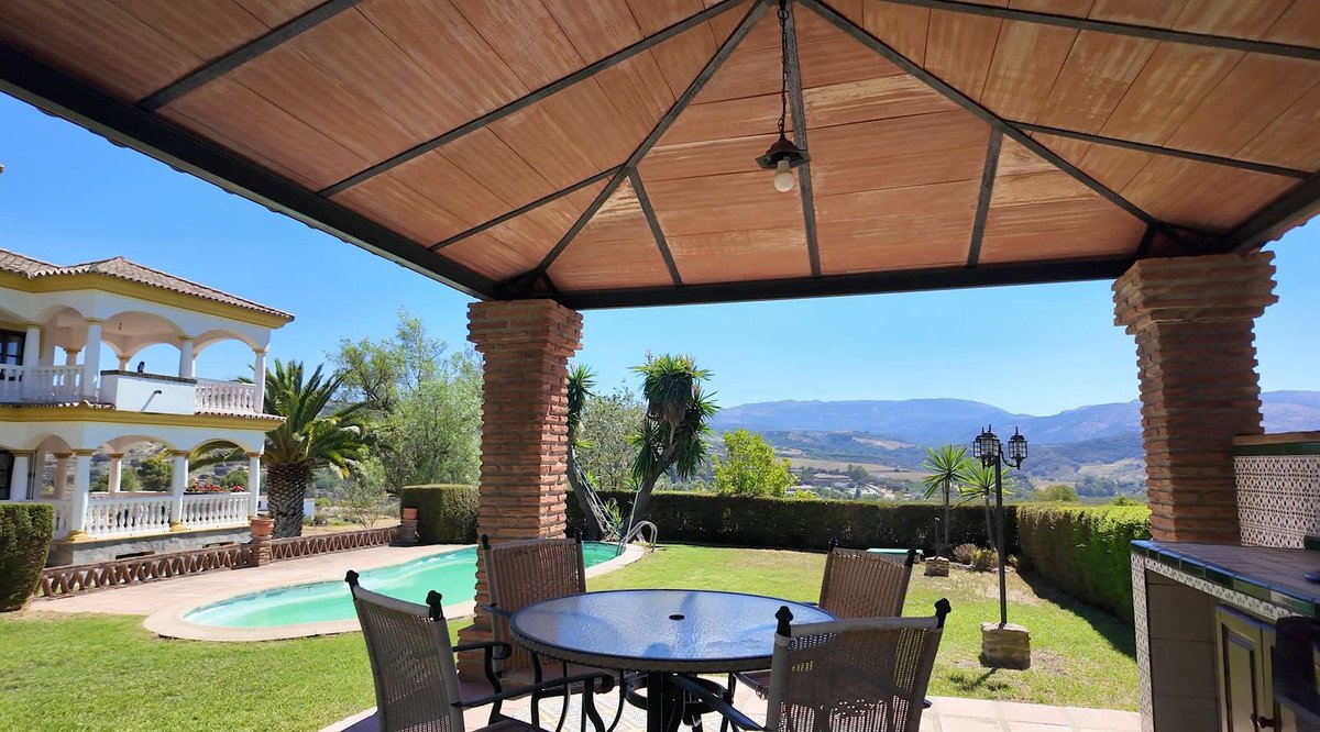 Covered patio with dining table overlooking a pool and distant hills. Features a large villa and palm trees.