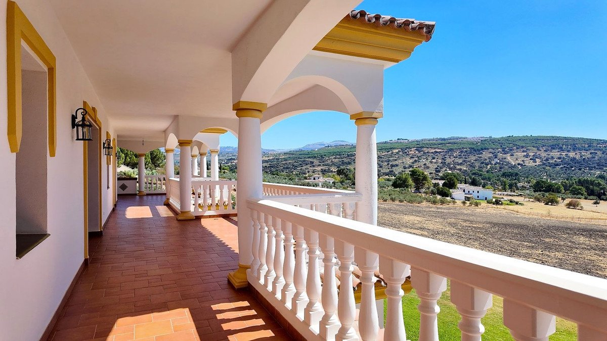 A covered terrace with white balustrades overlooks a picturesque landscape of rolling hills and distant mountains under a clear blue sky.