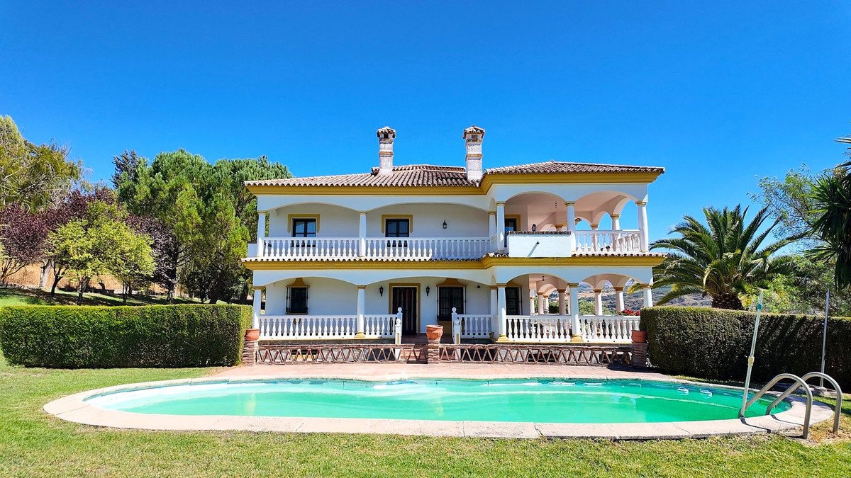 A large white villa with balconies and a swimming pool in front, surrounded by green lawn and trees under a clear blue sky.
