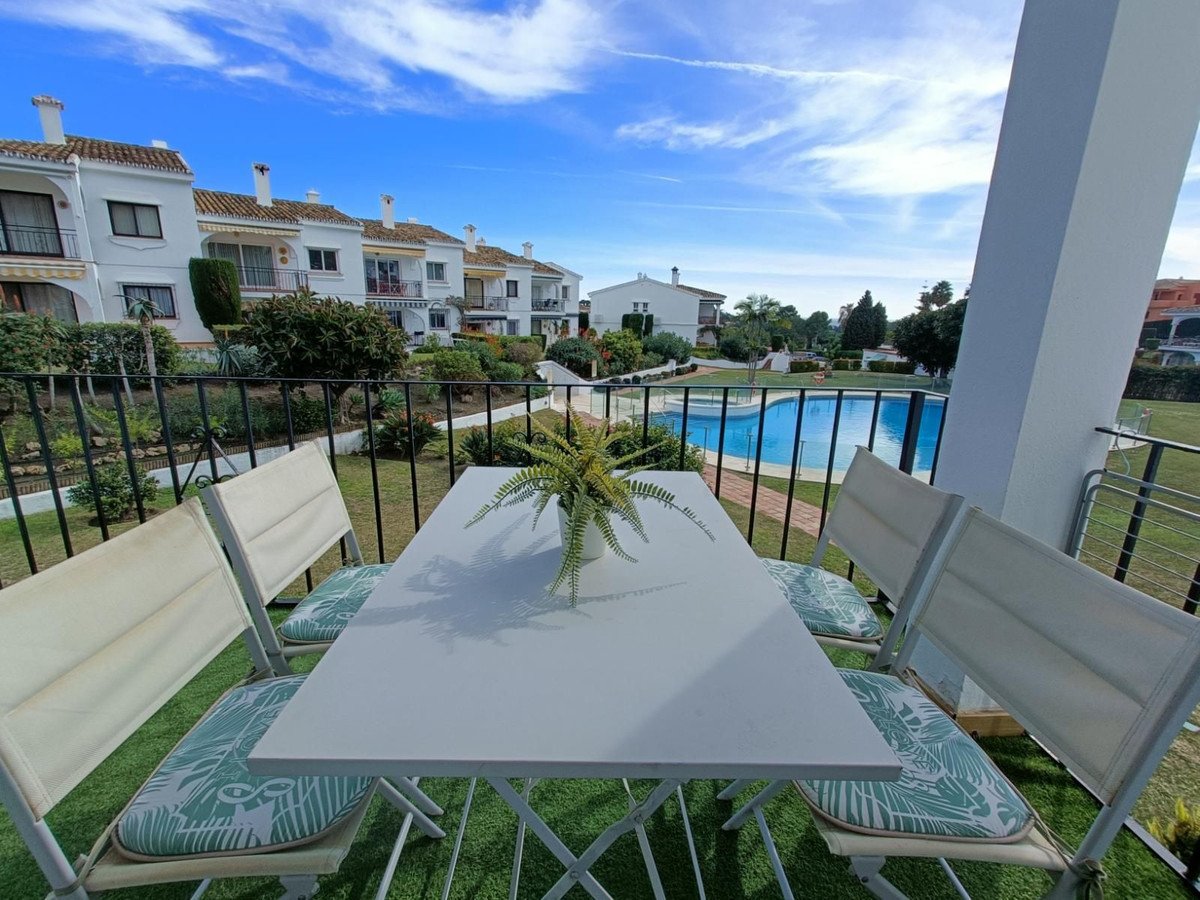 Balcony with table and chairs overlooking a swimming pool and manicured gardens, with white residential buildings in the background.