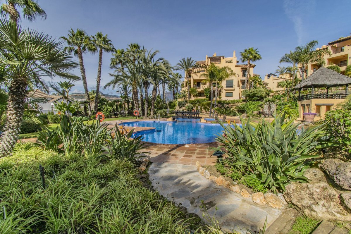 A resort complex with a swimming pool, palm trees, and lush tropical gardens under a clear blue sky.