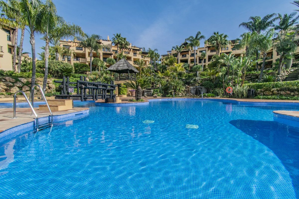 A large blue swimming pool with steps and palm trees, overlooking resort buildings and lush greenery.