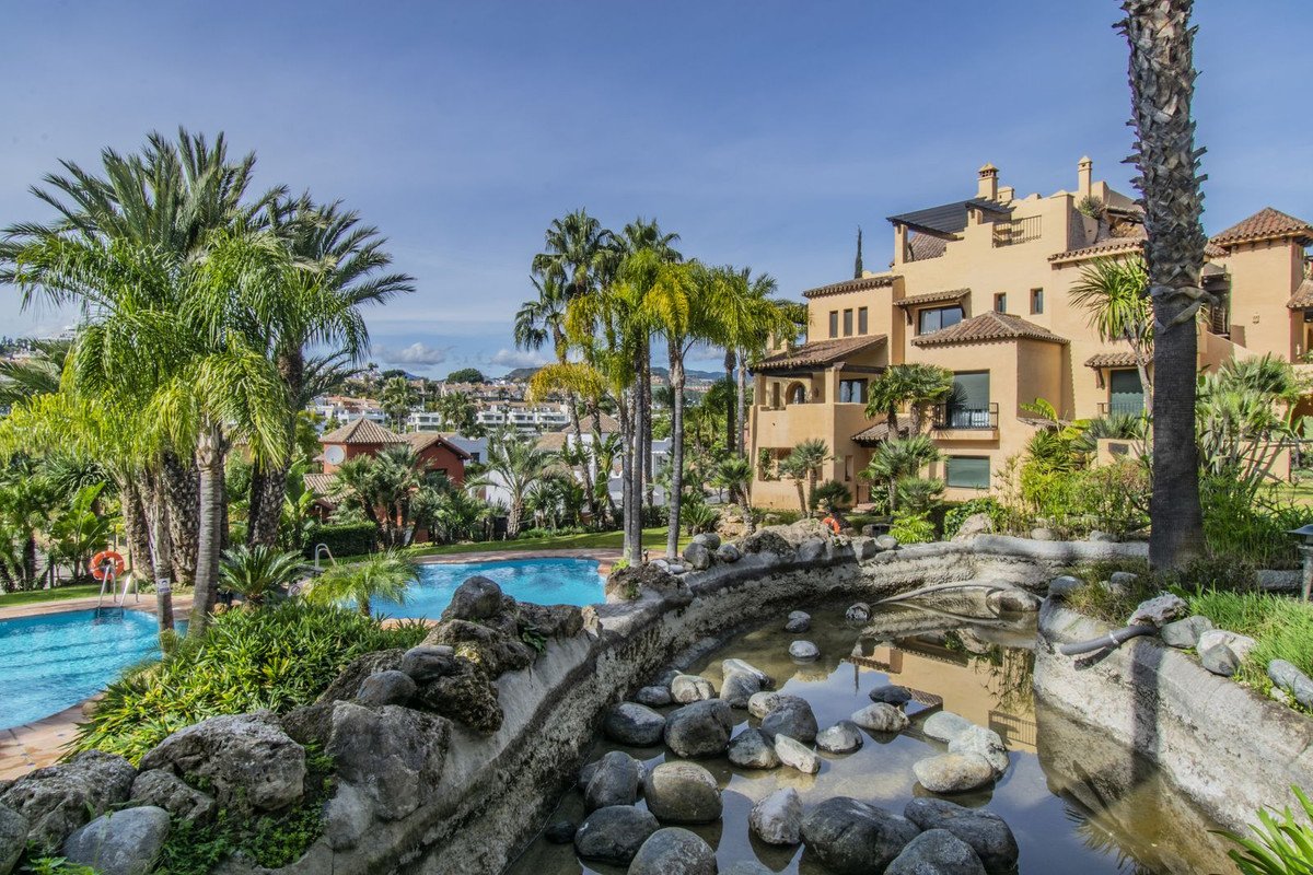 Apartment building with pools, palm trees, and a decorative water feature in a sunny resort setting.