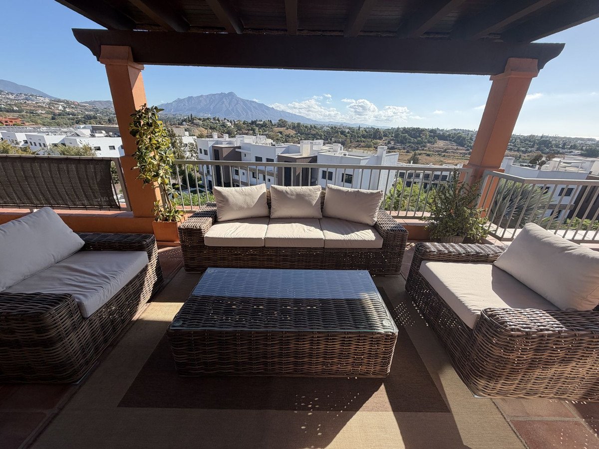 Outdoor terrace seating area with wicker furniture, overlooking buildings and a distant mountain range under a clear blue sky.