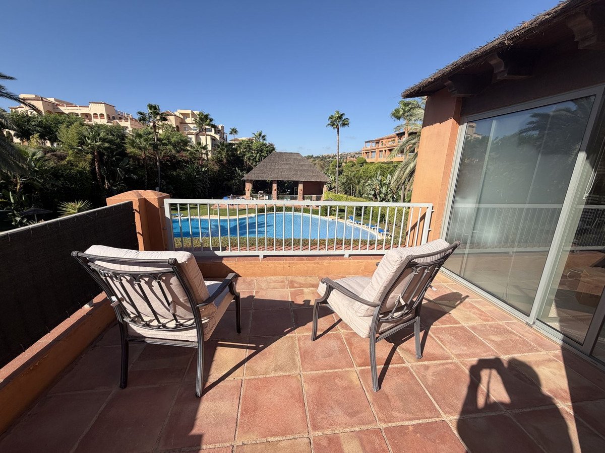 Terrace with two chairs overlooking a swimming pool and resort buildings, on a sunny day.