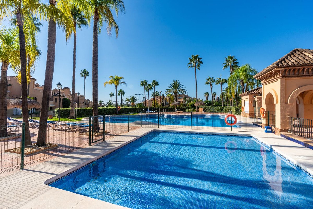 A resort-style outdoor swimming pool area with palm trees, loungers, and blue water under a clear sky.