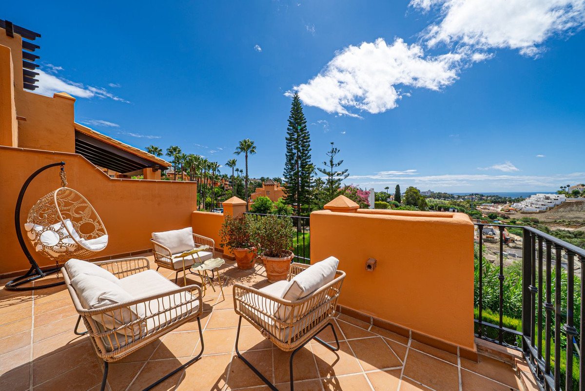 Outdoor terrace with wicker seating, a hanging chair, potted plants, and a view of the sea and coastline under a blue sky.