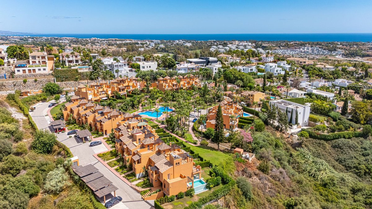 Aerial view of orange apartment buildings with pools and lush greenery, overlooking the sea under a clear blue sky.