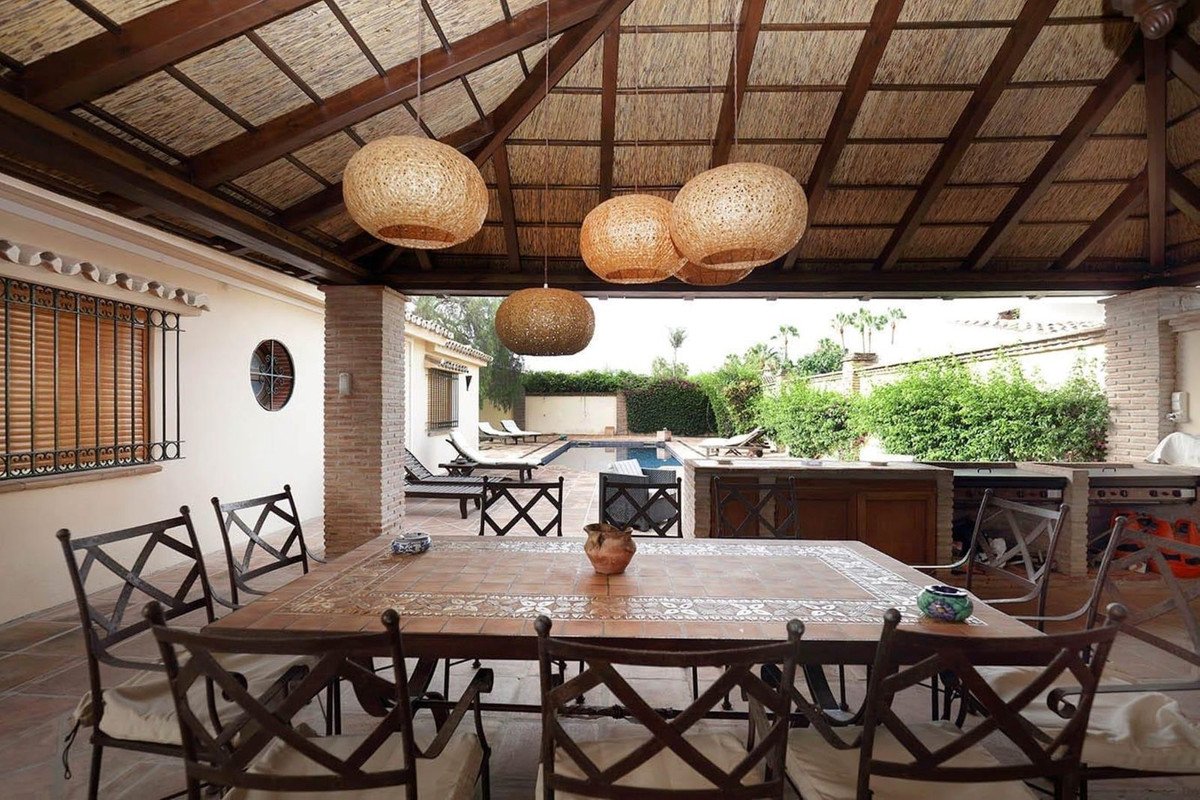 Covered patio with dining table, outdoor kitchen, and pool area with lush greenery and palm trees.