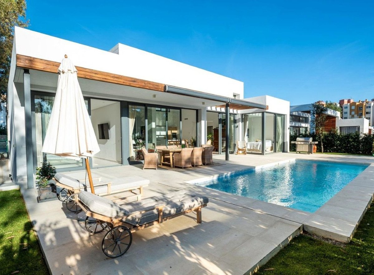 Modern villa with a swimming pool, outdoor dining area, lounge chairs, and a barbecue on a sunny day.