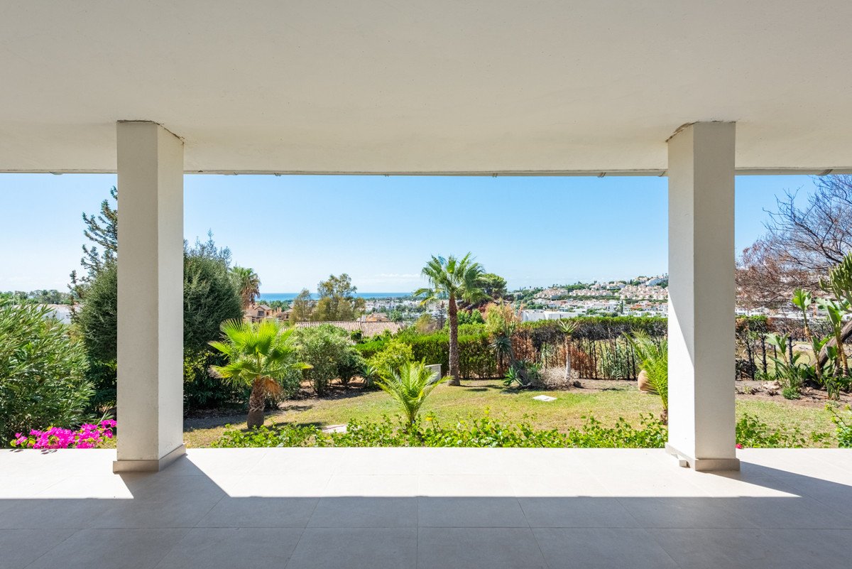 Veranda with white columns overlooking garden, palm trees, and distant sea view.