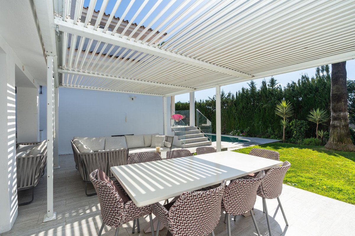 Outdoor patio with dining table, chairs, and sofa under a louvred roof, overlooking a swimming pool and garden.