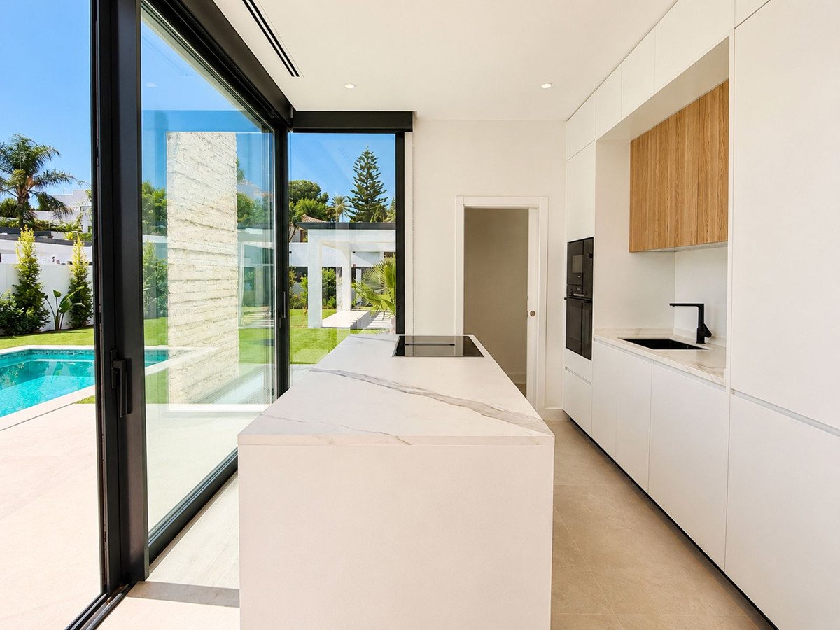 Modern white kitchen with island, black fixtures, and large sliding glass doors opening to a backyard pool and green lawn.