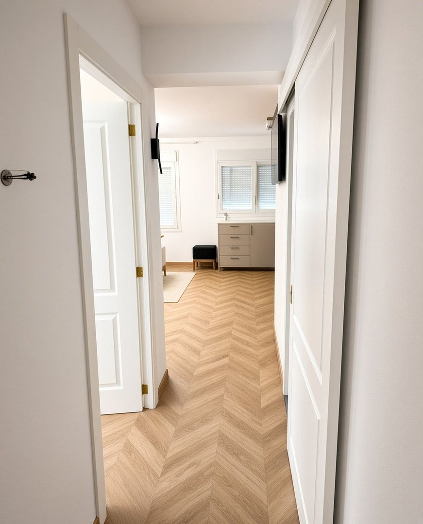 A modern hallway with herringbone wood flooring, leading to a room with a TV and windows.