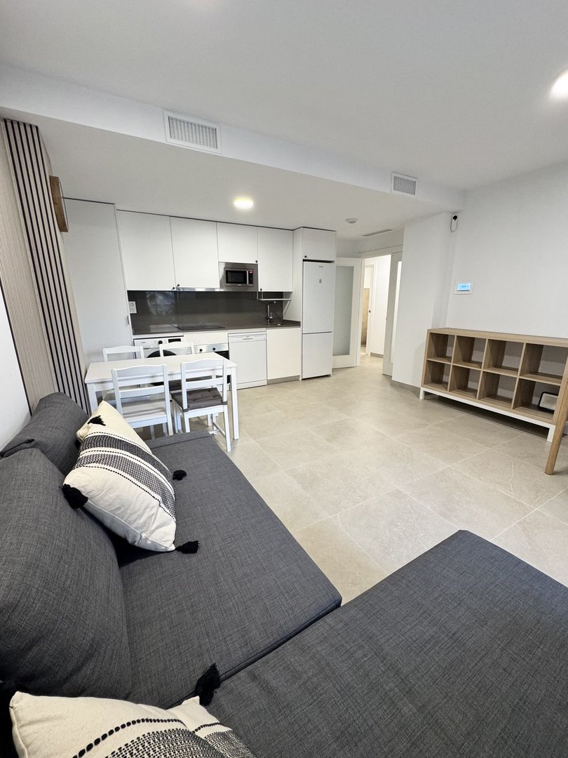 A modern open-plan living area with a sofa and a fully equipped kitchen. Modern furnishings and light grey tiled floor.